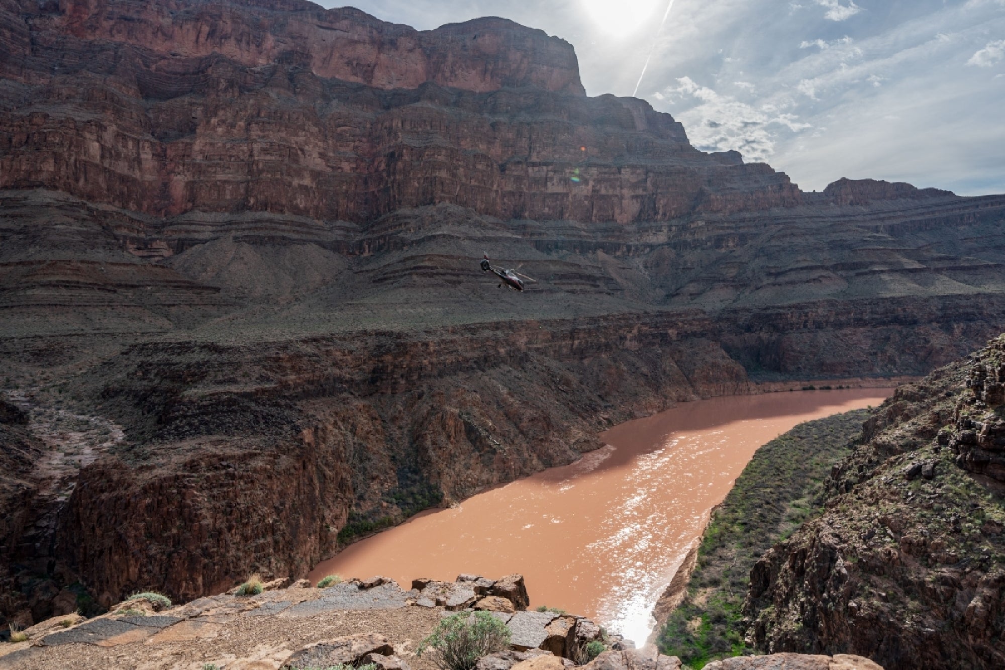 Helicopter soaring above river beside rugged canyon walls.