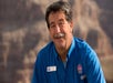 A man with gray hair and a mustache wearing a blue shirt with a name tag stands outdoors with rocky cliffs in the background.