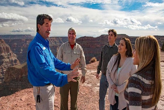 A tour guide speaks to four people standing on a rocky overlook with a canyon landscape and cloudy sky in the background.