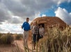 A group of people stand and talk outside a traditional round, earth-covered structure in a desert landscape under a partly cloudy sky.