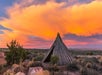 A wooden teepee stands in a desert landscape with a vibrant orange and pink sunset sky.
