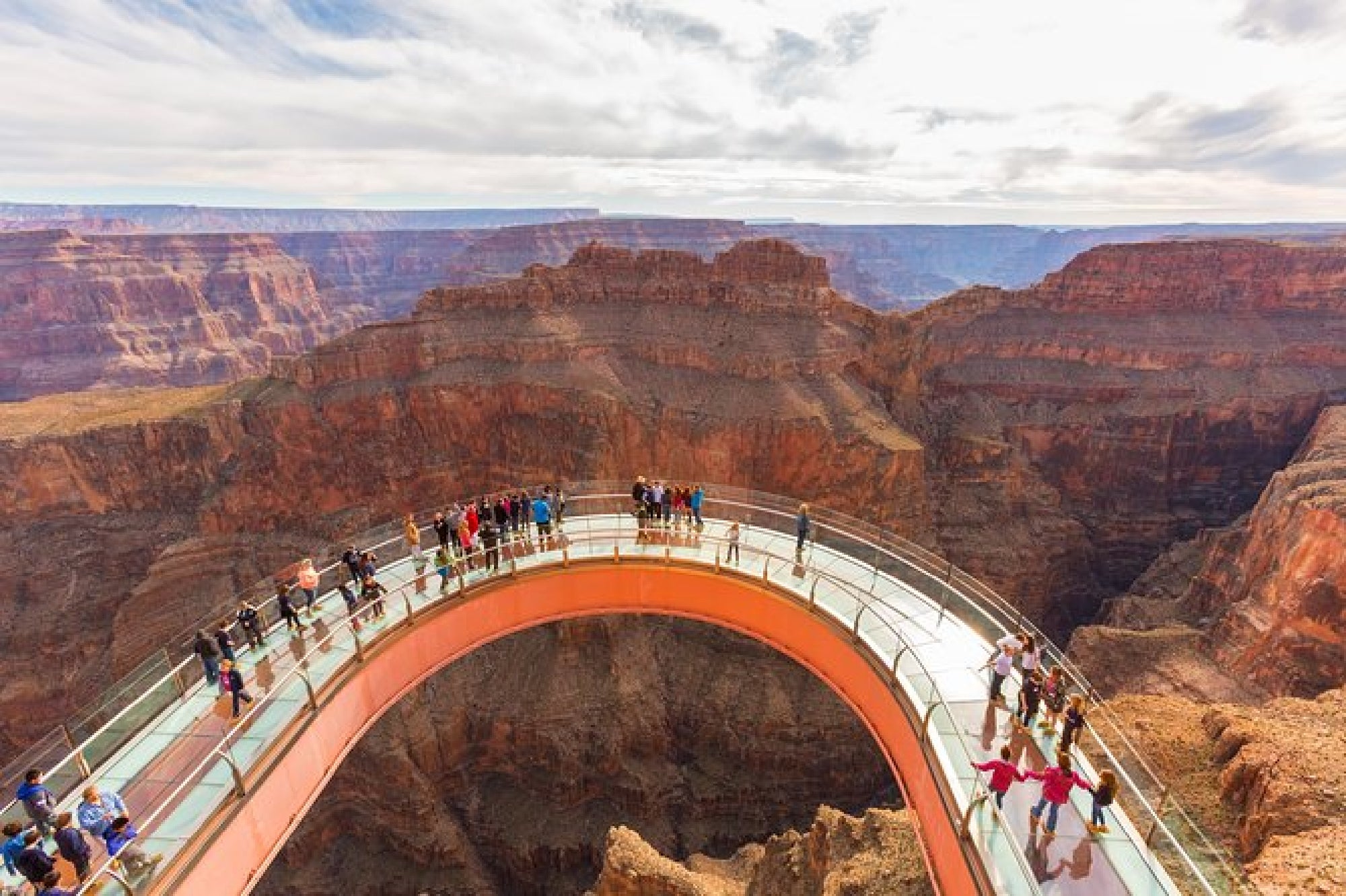 People stand on the glass-bottomed Skywalk overlooking a deep canyon at the Grand Canyon, with rocky cliffs and a cloudy sky in the background.