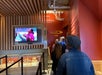 People stand in line inside a building, watching a screen with a video, under a sign labeled "Skywalk" with an arrow pointing right.