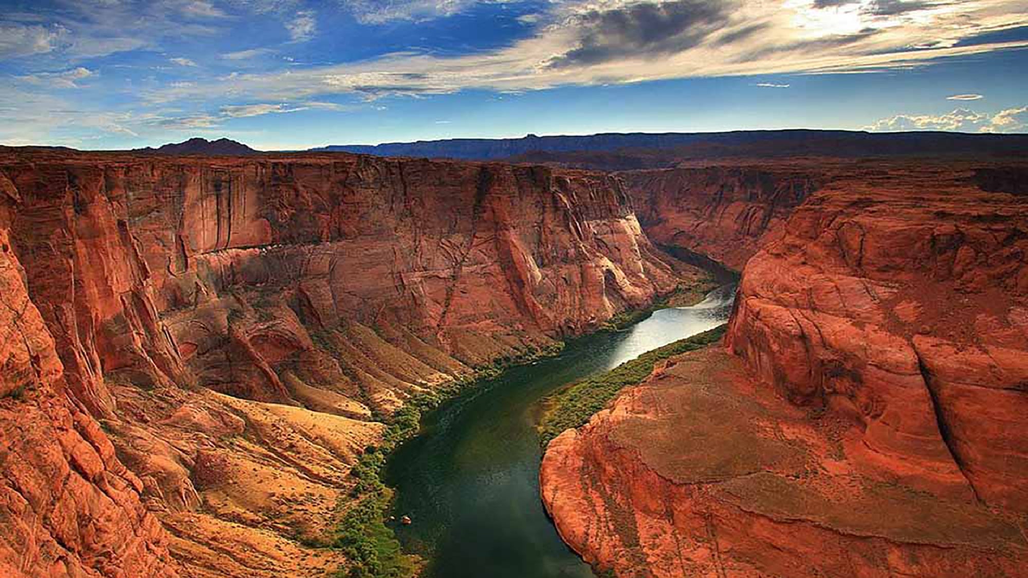 A river curves between steep red rock cliffs under a partly cloudy sky, showcasing a dramatic landscape.