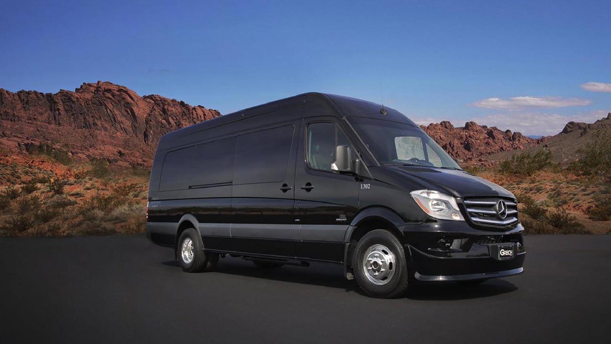 A black Mercedes-Benz Sprinter van parked on an asphalt road with rocky desert mountains in the background under a partly cloudy sky.