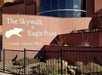 Sign reading "The Skywalk at Eagle Point — Grand Canyon West" with a bird silhouette, mounted on a curved wall behind a black metal fence with desert plants.