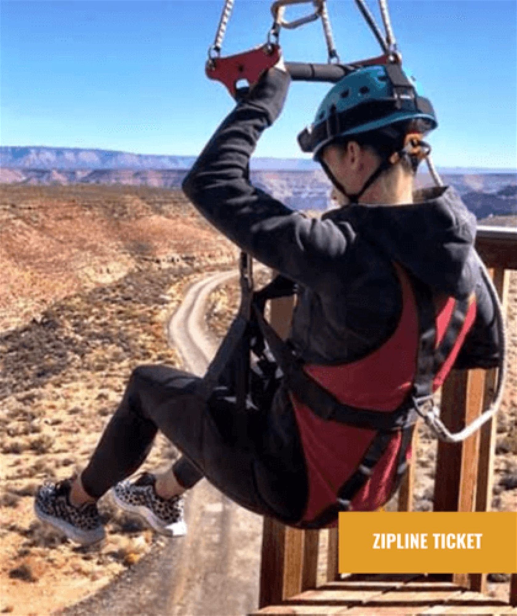 Person wearing helmet and harness starts a zipline ride over a desert landscape with a winding dirt road visible below.