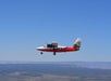 A small red and white propeller airplane with a tour company logo flies above a vast landscape under a clear blue sky.