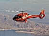 A red tour helicopter with white markings flies over a rugged, mountainous landscape with a body of water below.