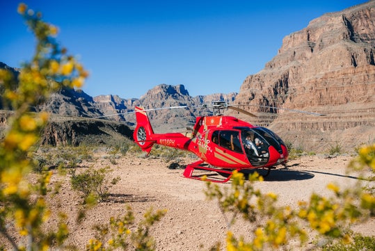 A red helicopter is parked on a rocky, desert terrain with mountains in the background and some yellow-flowered shrubs in the foreground.