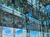 A mirrored observation deck reflects people and city buildings, creating an optical illusion. The sky and clouds are visible through large glass windows.