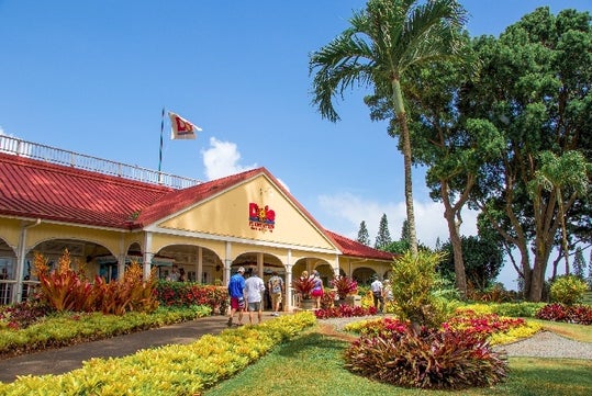 The front entrance to the Dole Pineapple Plantation with beautiful tropical landscaping and people walking into the building.