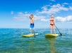 A man and woman in swim suits standing on yellow paddle boards in the ocean on a sunny day with clouds in the sky.