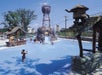 Children play in a water park with a wooden water slide and structures. A large bucket pours water from above in a sunny setting.