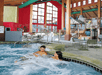 Two people relax in a large indoor hot tub with bubbling water. The room features high ceilings, tall windows, and a colorful area with water play structures in the background.
