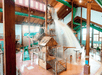 Indoor water park with a large wooden-themed play structure, slides, and a big bucket pouring water, surrounded by pools and high ceilings.