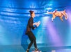 A person in black pants and a blazer plays disc with a dog performing an acrobatic leap on a stage with a blue backdrop.