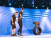 Two people on a stage with two dogs; one dog stands on stacked tires, and the other sits inside them. Blue backdrop with lights.