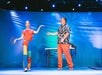Two people on stage, one wearing colorful clothing and gesturing, the other in a patterned shirt and orange pants. A table and a blue backdrop are behind them.