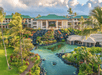 A luxurious coastal resort surrounded by lush gardens and a pond, with a main building featuring a green roof and tropical landscaping under a partly cloudy sky.