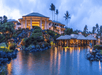 Luxurious resort at dusk with illuminated buildings, palm trees, and a tranquil water reflection in a tropical setting.