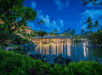 Luxurious tropical villa at night, with illuminated windows and reflections on a calm pond, surrounded by lush greenery and palm trees under a deep blue sky.
