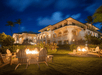 Outdoor seating with fire pits in front of a large, well-lit building at night, surrounded by tropical plants and palm trees.