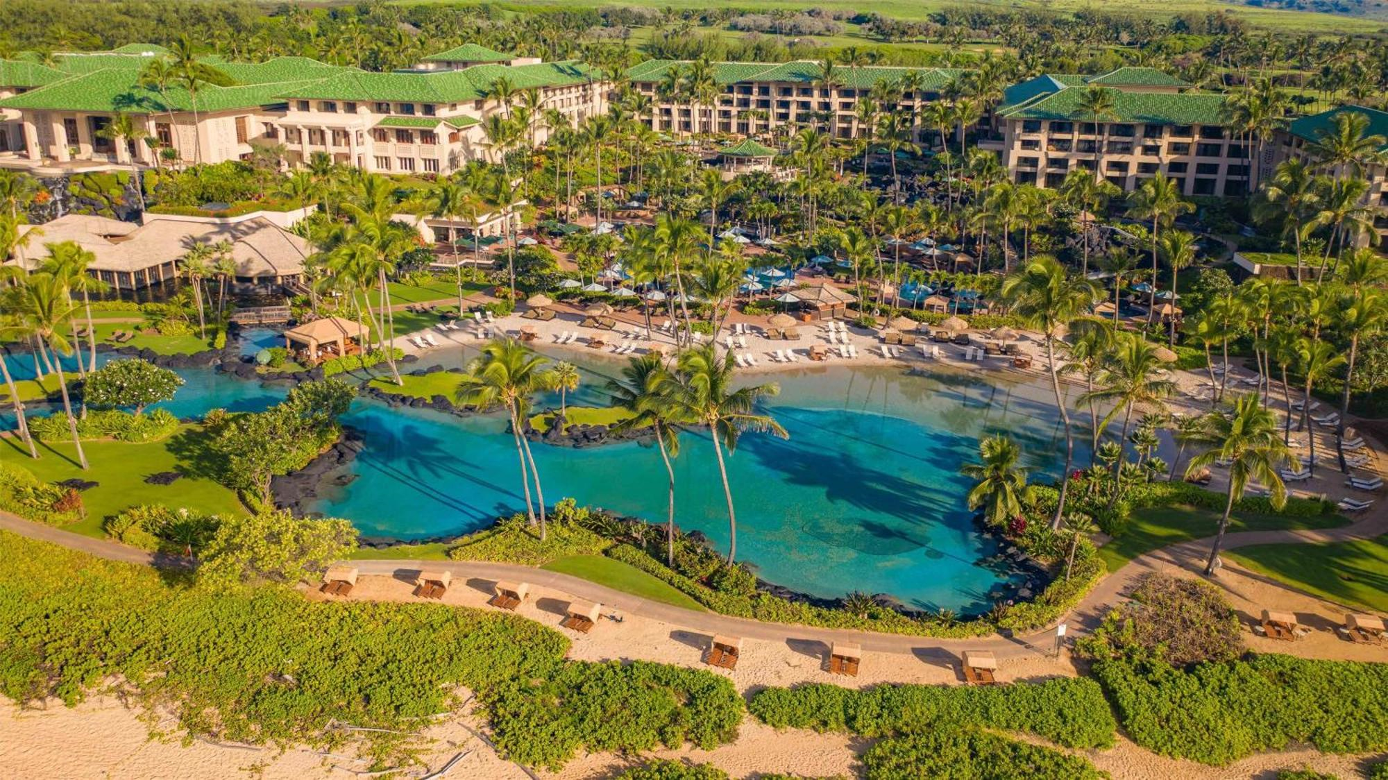 Aerial view of a large tropical resort with green-roofed buildings, palm trees, multiple swimming pools, lounge chairs, and landscaped gardens.