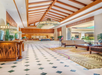 Spacious hotel lobby with high wooden ceiling, large light fixtures, tiled floor, indoor plants, and seating area with sofas and wooden tables.