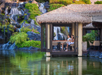 Two people sit at a table in a thatched-roof pavilion over water, with lush greenery and small waterfalls in the background.