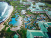 Aerial view of a large beachfront resort with multiple pools, green rooftops, landscaped gardens, and direct access to the ocean.