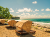 Wooden sunbeds with canopy covers are arranged on a sandy beach near the ocean, with rocky shoreline and clear skies in the background.