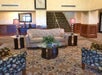 Hotel lobby with a beige sofa, patterned chairs, glass-topped tables, lamps, a decorative plant, and a staircase. Large clock on the wall above the couch.