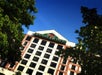 Exterior view of a multi-story hotel building with a white and brown facade, framed by green leafy trees, under a clear blue sky.