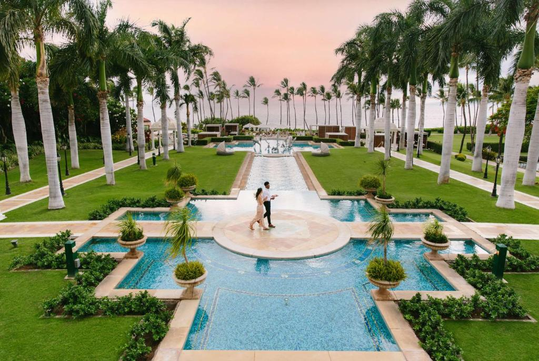 A couple walks along a pathway surrounded by pools and palm trees in a landscaped garden with a sunset view.