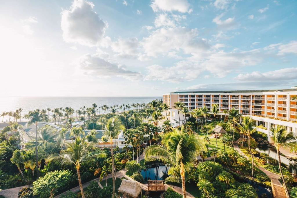 Aerial view of a seaside resort with a large building on the right, palm trees, and landscaped gardens, overlooking the ocean under a partly cloudy sky.