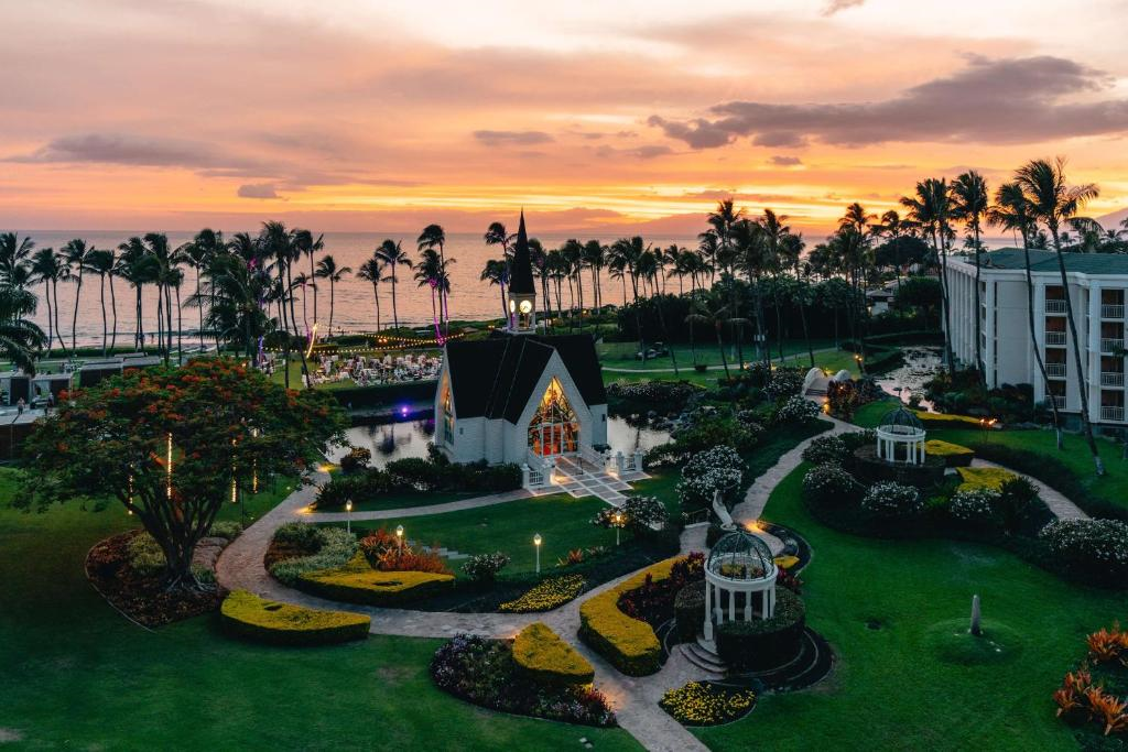 Aerial view of a beachfront resort at sunset, featuring a small chapel, gardens, and palm trees.