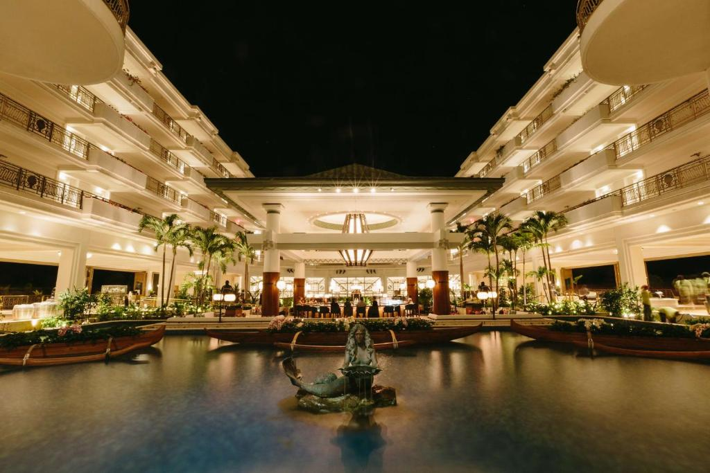A hotel with lit balconies and palm trees surrounds a central courtyard and pool with a statue, under a nighttime sky.