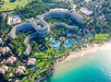 Aerial view of a large beachfront resort with multiple buildings, swimming pools, and lush greenery, situated along a sandy beach and turquoise ocean.