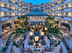 Spacious hotel atrium with multiple floors, central bar area, seating arrangements, surrounding palm trees, and warm lighting.