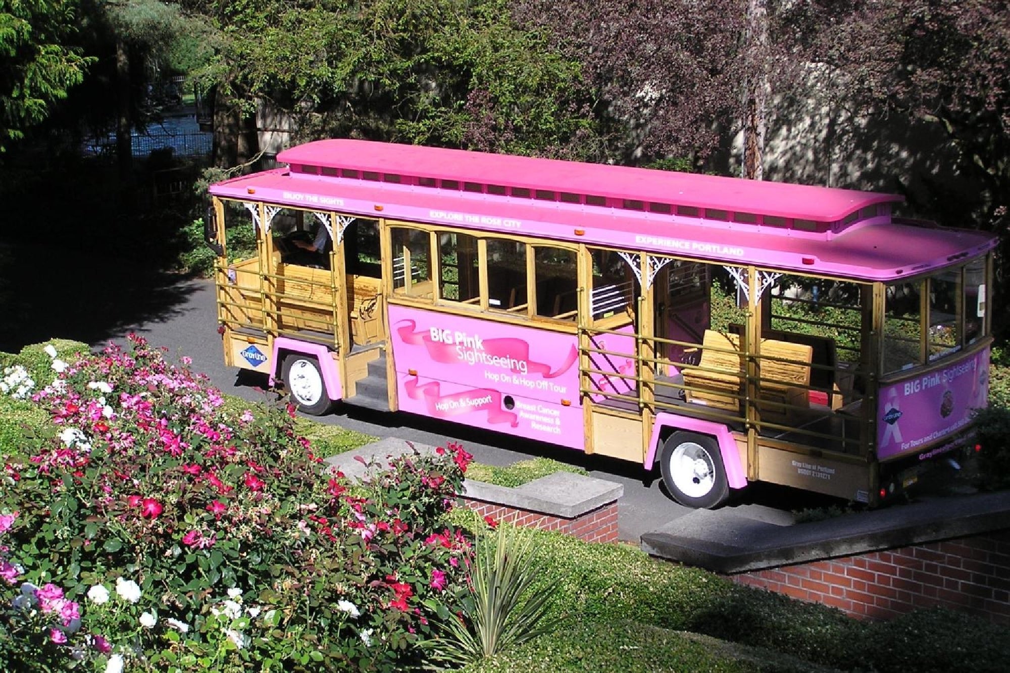 A bright pink tour trolley stands on a tree-lined street, surrounded by colorful flowers and greenery.