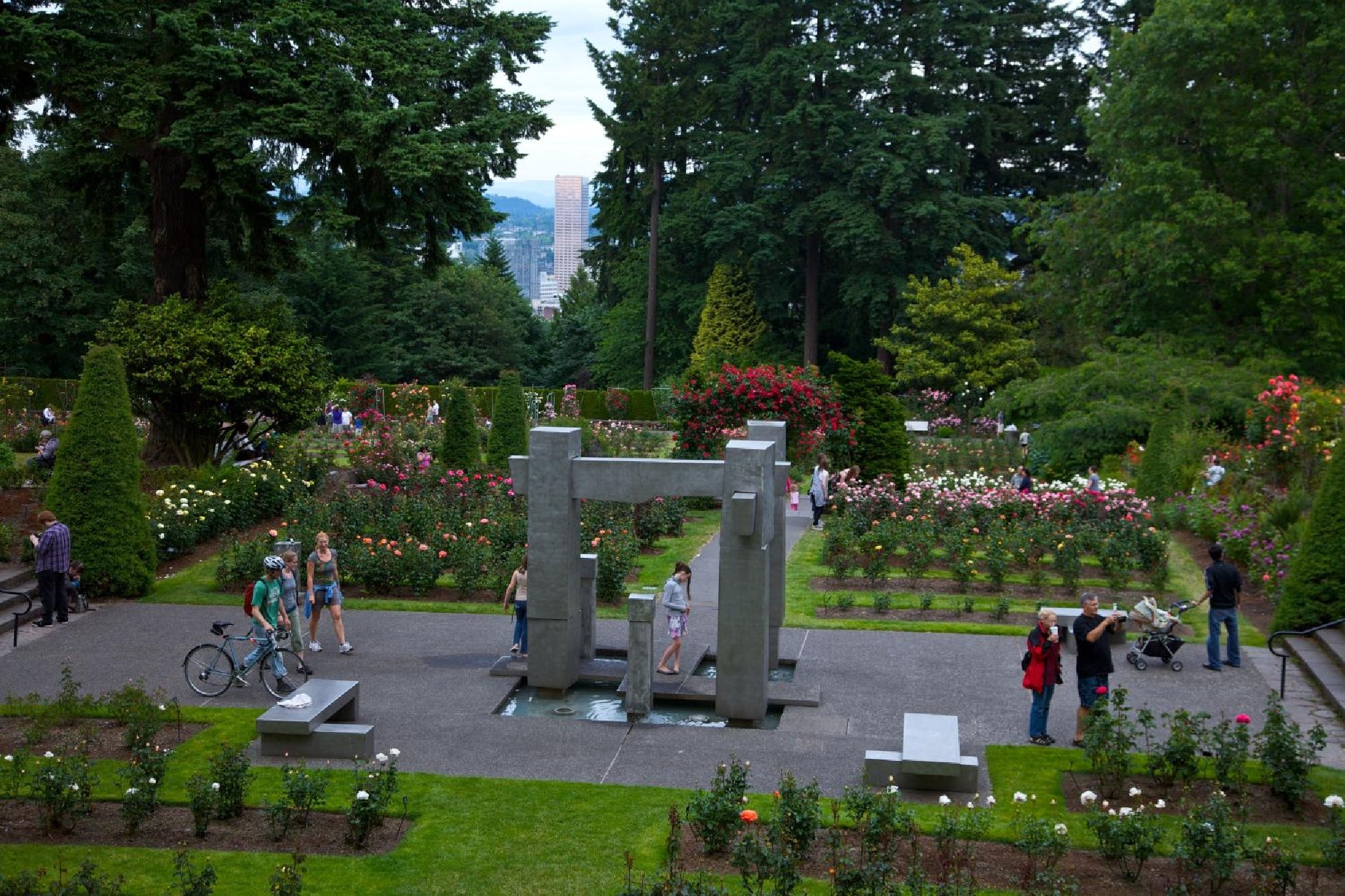 A formal garden scene with people walking and relaxing among rose bushes, featuring a large, abstract stone sculpture in the center. Trees and a city skyline are visible in the background.