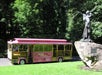 A pink sightseeing trolley is parked near a statue of a woman and child in a green, wooded area.