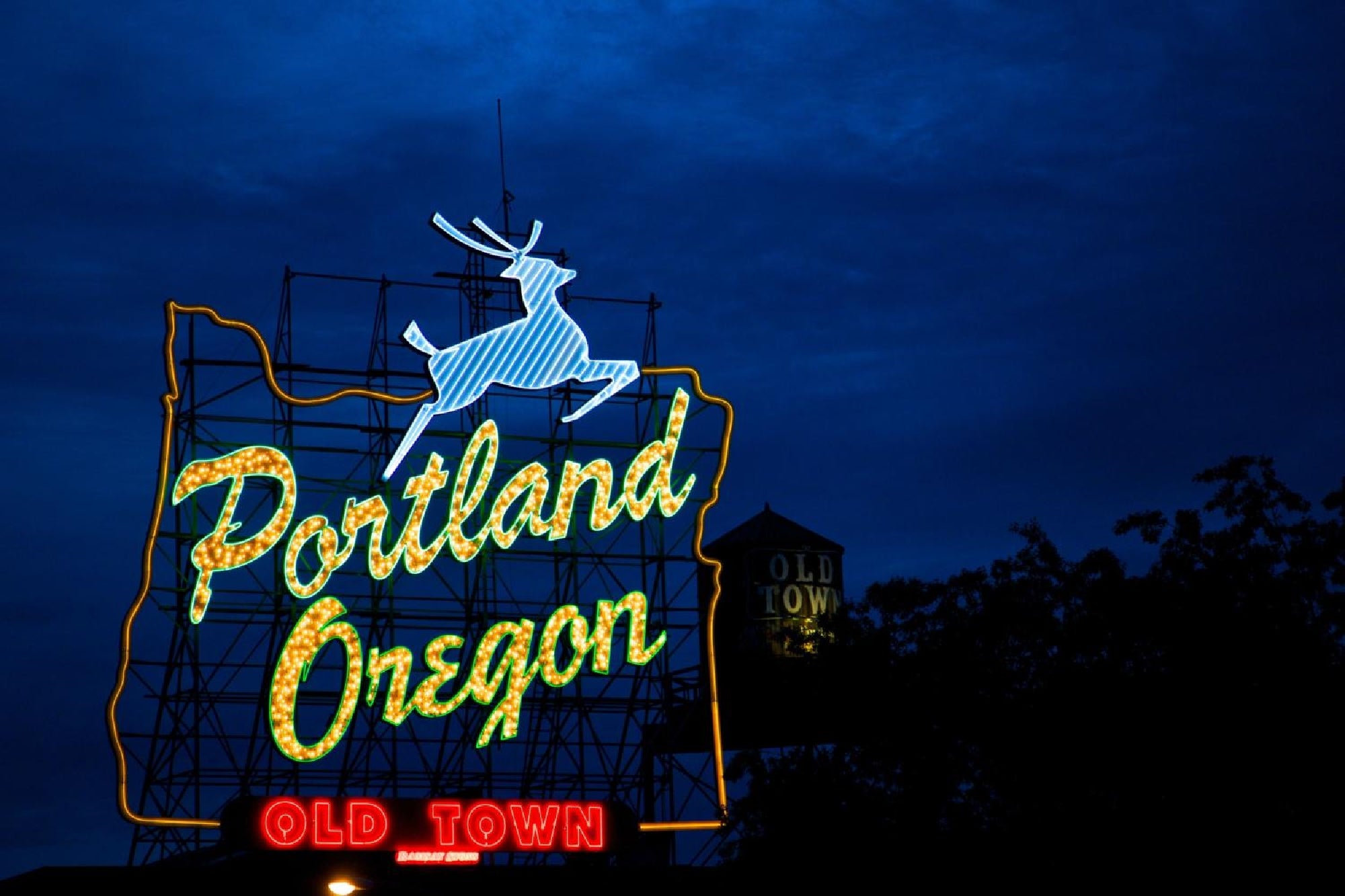 Neon sign in Portland, Oregon, featuring a leaping deer and outlined state shape, with "Portland Oregon" and "Old Town" text, against a dark blue sky.