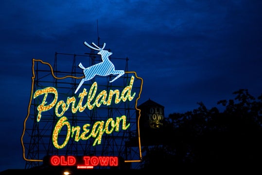 Neon sign in Portland, Oregon, featuring a leaping deer and outlined state shape, with "Portland Oregon" and "Old Town" text, against a dark blue sky.