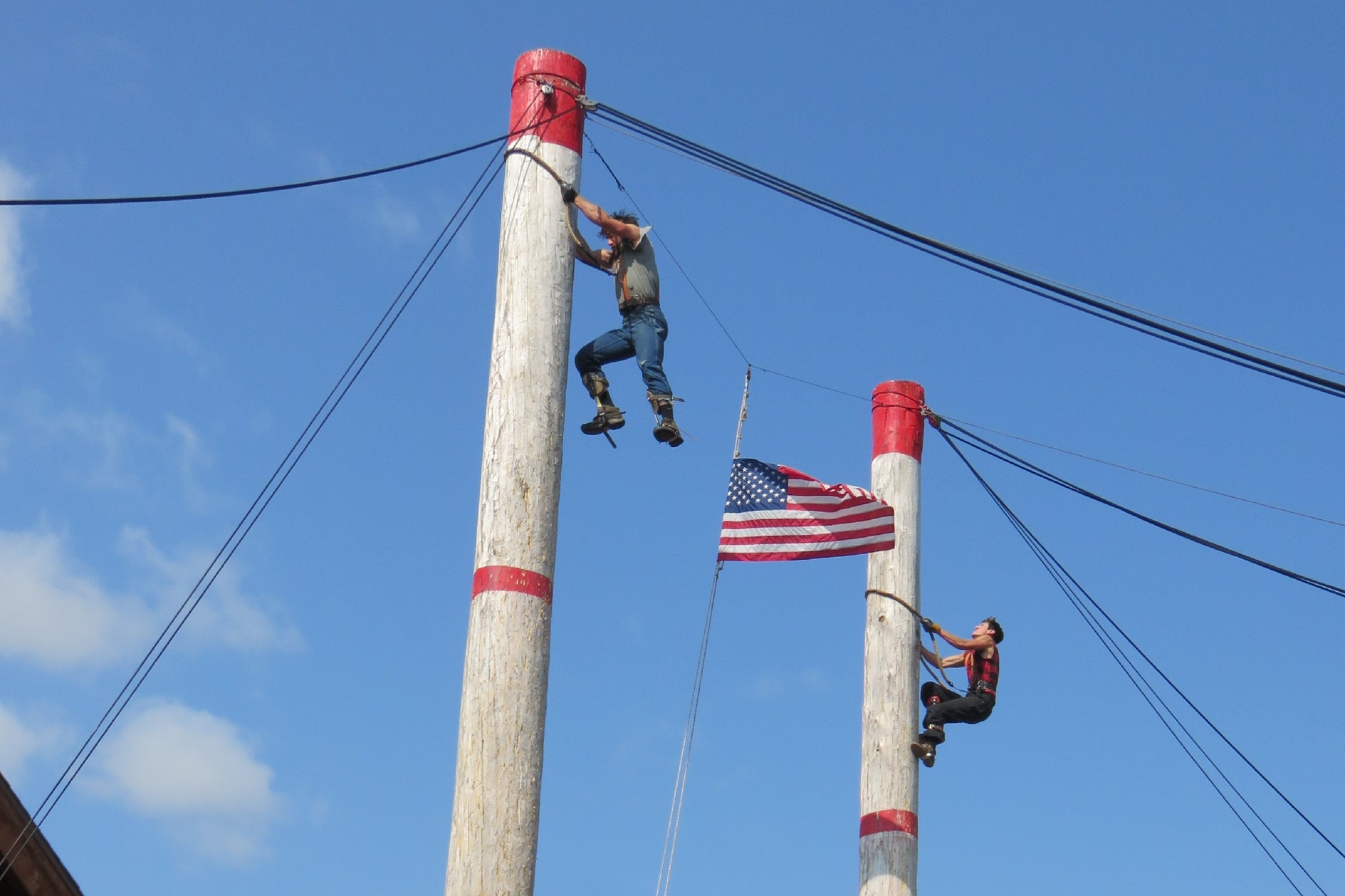 Two people climb tall wooden poles with ropes, while an American flag waves in the background against a clear blue sky.