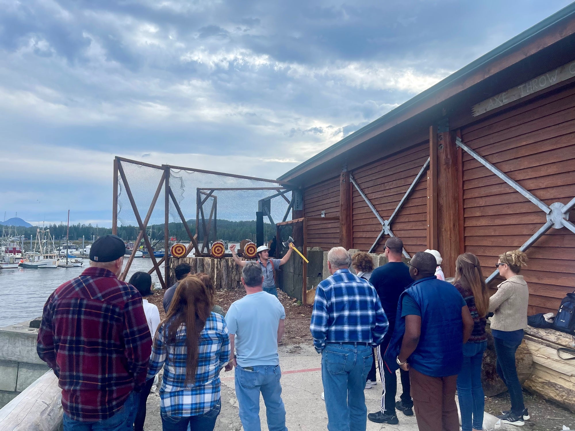 A group of people stand outside near a wooden building, watching axe throwing at targets set up by the water.