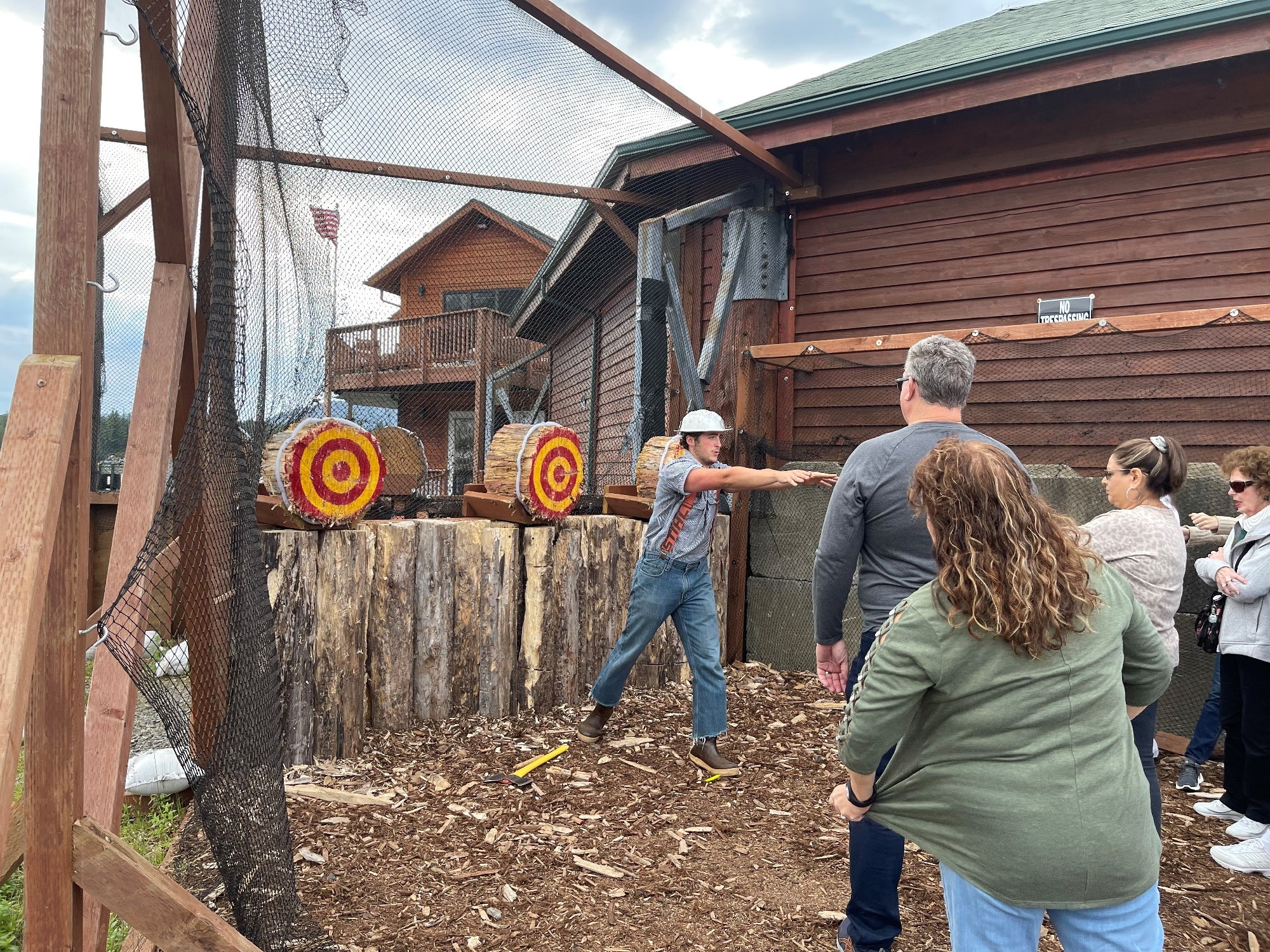 A man throws an axe at a target in an outdoor axe-throwing area while a small group of people watch.