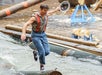 A man wearing suspenders and jeans balances on a floating log in water while holding a hat, participating in a log-rolling event.