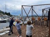 Three people throw axes at wooden targets in an outdoor axe-throwing area by a marina, with boats and buildings visible in the background.